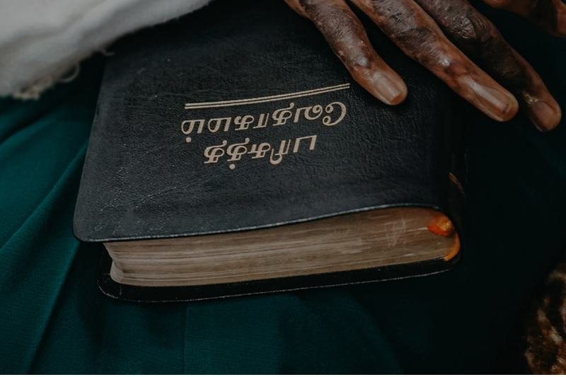 A hand resting on a worn, gilt-lettered Bible printed in Tamil script.